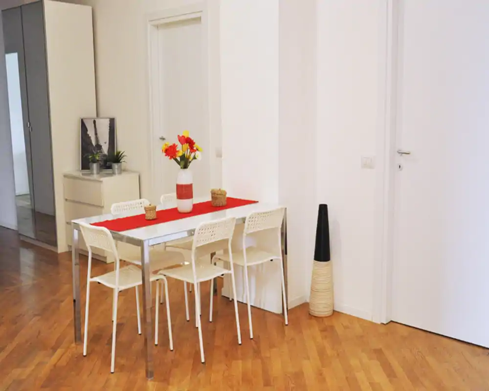 Área de comedor moderna y luminosa con mesa blanca, seis sillas, camino de mesa rojo y un jarrón decorativo sobre suelo de madera cálida — espacio sencillo e invitante para comer.