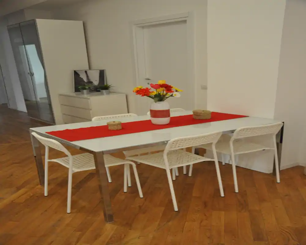 Zona de comedor moderna y luminosa con mesa de cristal para seis, camino de mesa rojo y un jarrón de flores sobre suelo de madera cálida.