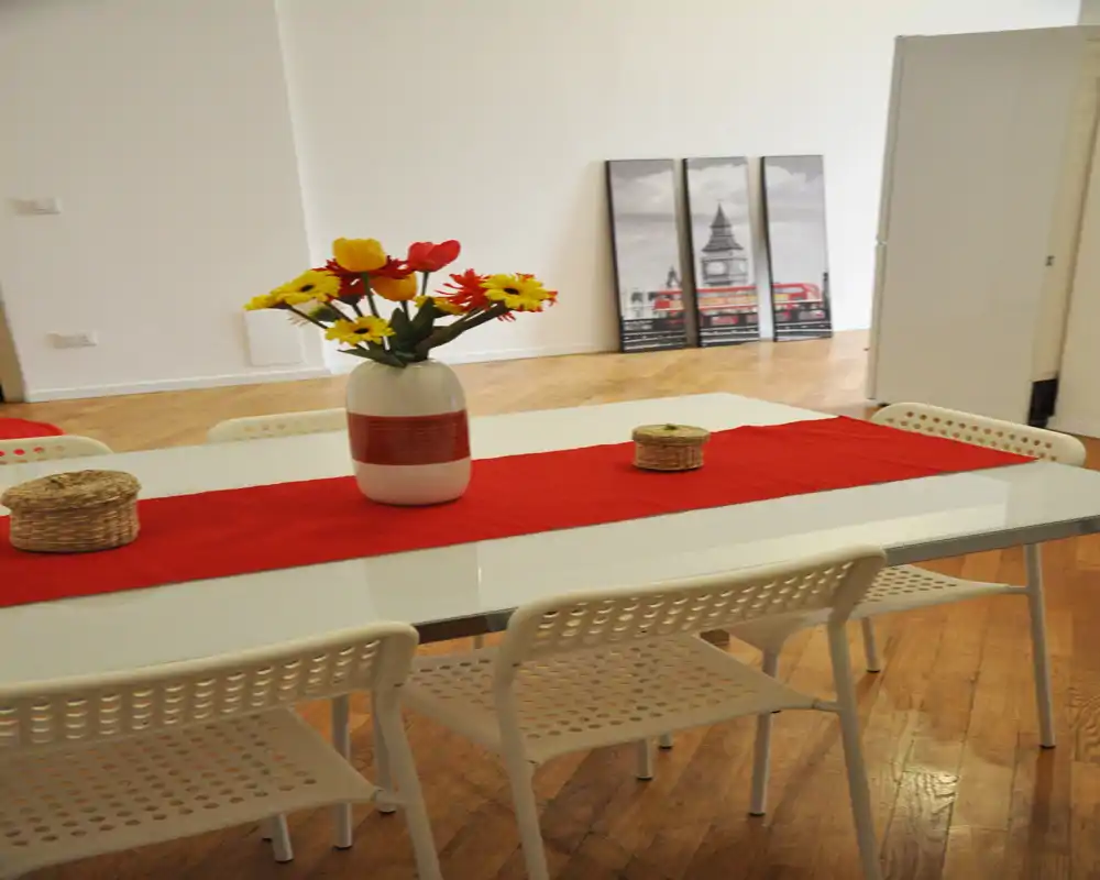 Zona de comedor luminosa y decorada con mesa blanca, varias sillas y un jarrón de flores en un corredor rojo — moderna y acogedora para comer en compañía.