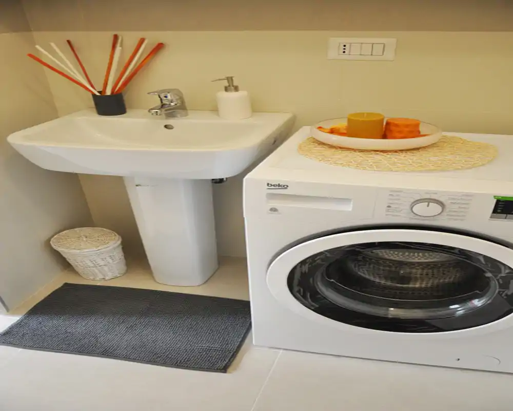 Baño compacto con rincón de lavandería: lavabo de pedestal y lavadora de carga frontal, decorado con una alfombra y objetos sobre la lavadora.
