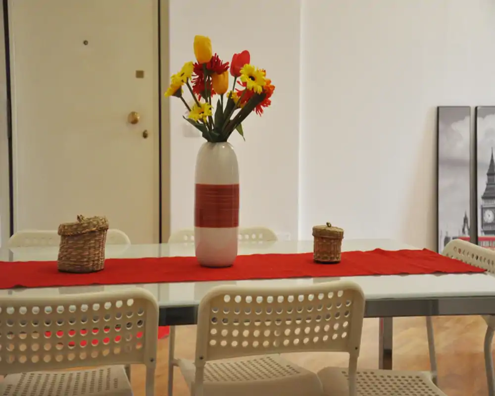 Zona de comedor luminosa y arreglada con mesa de cristal, sillas blancas perforadas y un jarrón de flores coloridas sobre una banda roja — ideal para el anuncio.