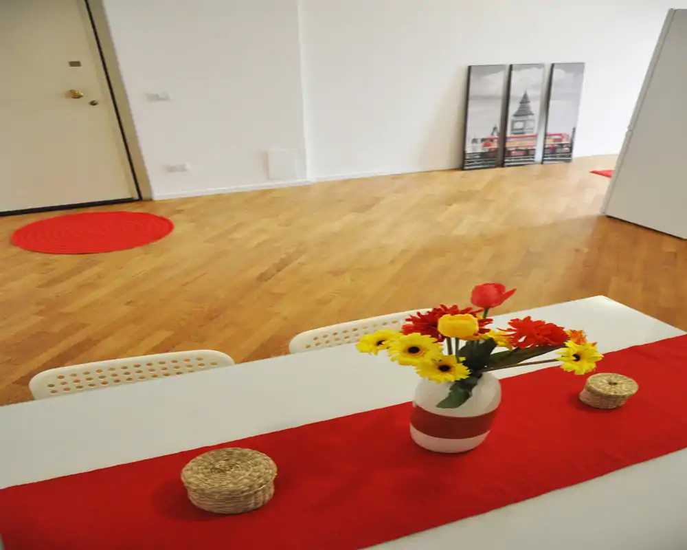 Zona de comedor luminosa y sencilla con mesa blanca, camino de mesa rojo y jarrón de flores, sobre suelos de madera cálida — ideal para mostrar un comedor compacto.