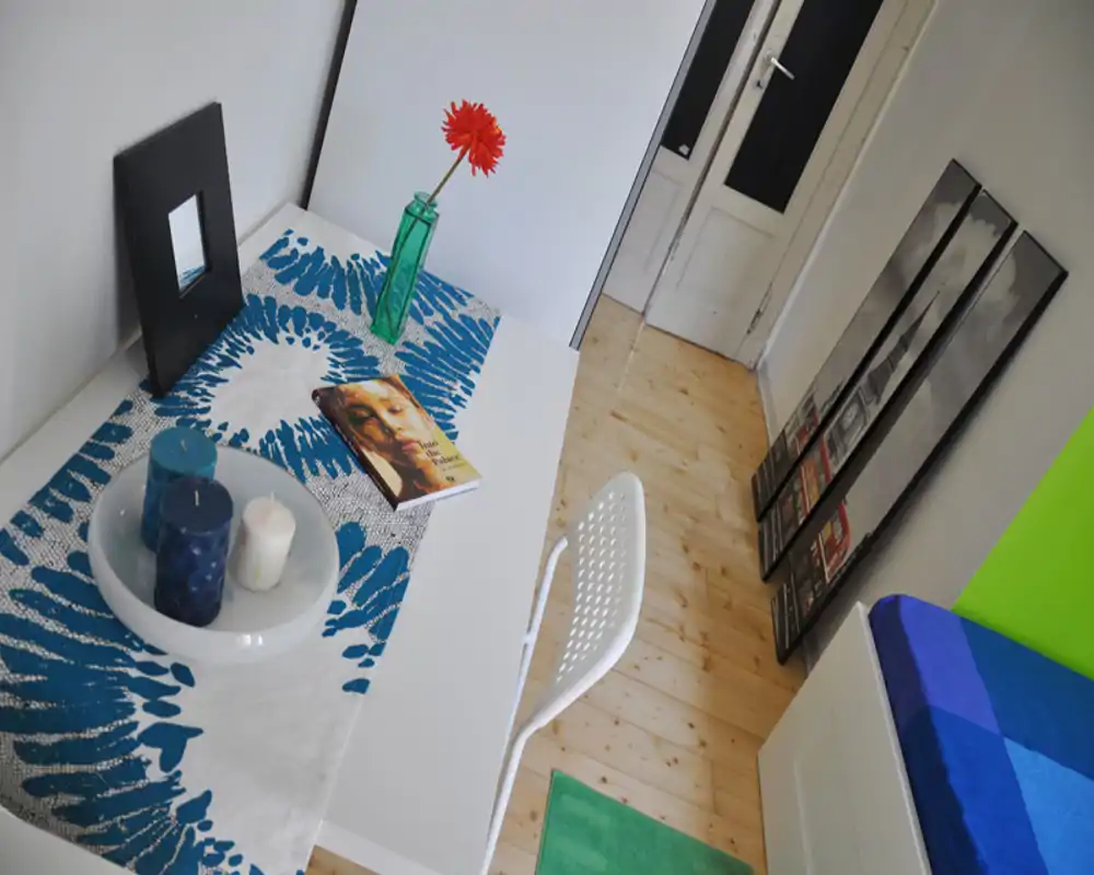Rincón de dormitorio luminoso y moderno con escritorio blanco y silla junto a la cama azul y verde. Detalles decorativos: velas, un libro y una flor roja en un jarrón de cristal sobre un camino de mesa estampado.