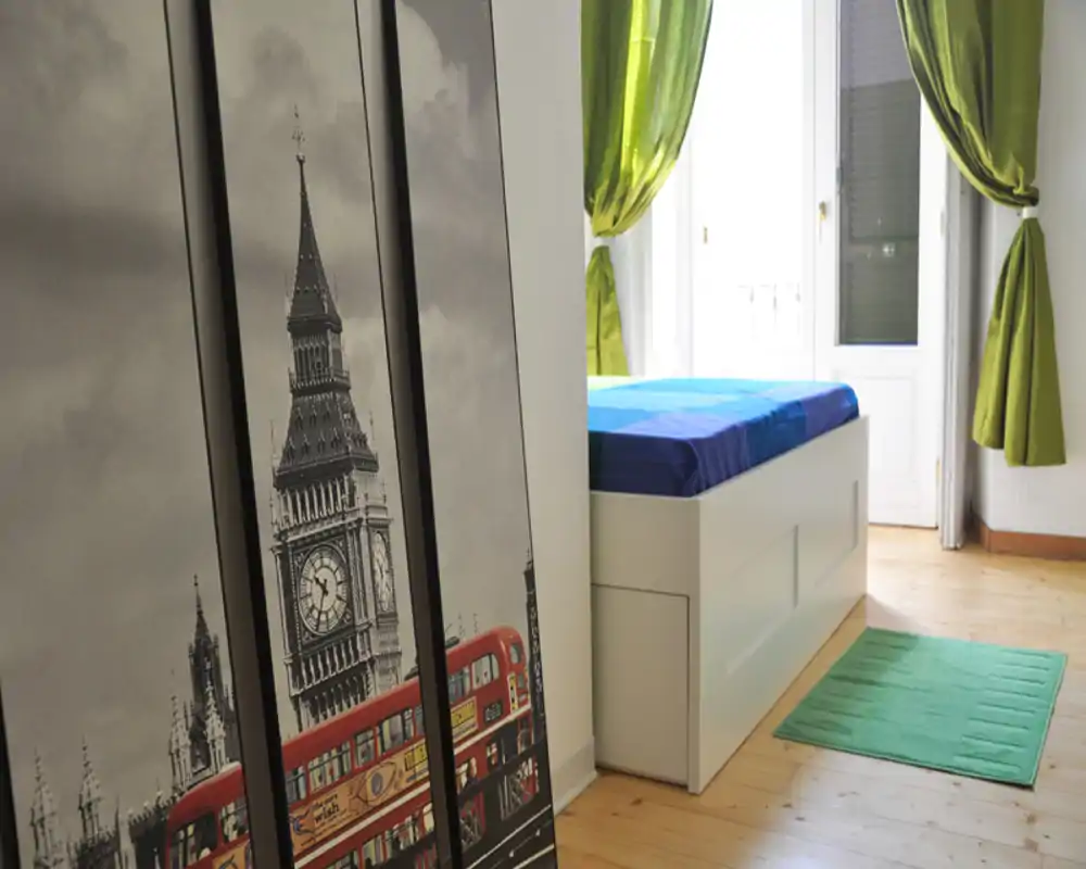 Dormitorio individual luminoso con cama con almacenaje blanca, cortinas verdes y suelo de madera. Estilo limpio y moderno, con una alfombra pequeña y una gran ventana que deja entrar mucha luz.