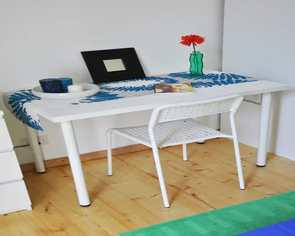 Rincón de trabajo luminoso y minimalista con mesa blanca y silla perforada, decorado con un camino de mesa, velas y una flor roja en un jarrón verde.
