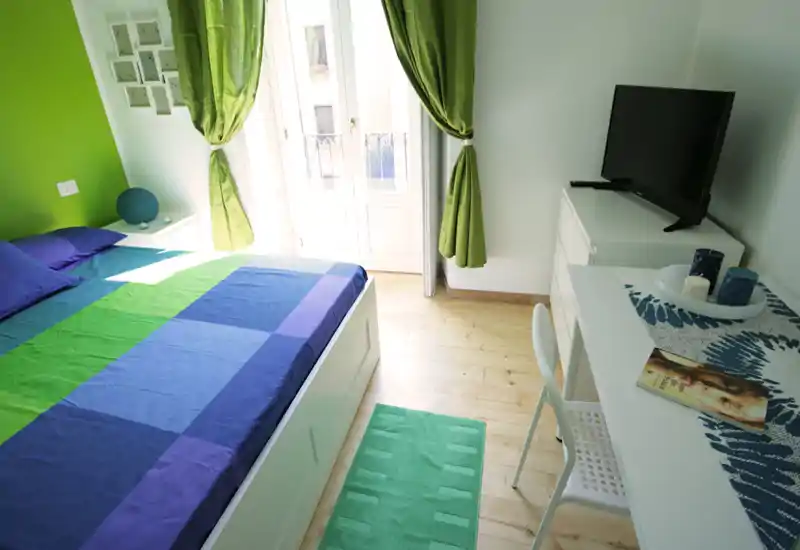Dormitorio moderno y luminoso con una cama grande con ropa de cama verde y azul, un escritorio con TV y una puerta al balcón que aporta abundante luz natural.