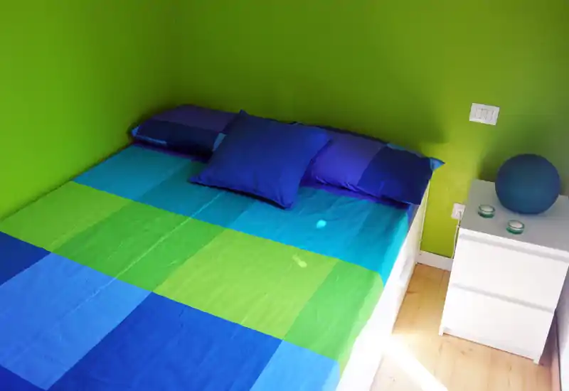 Dormitorio luminoso y colorido con cama doble vestida en tonos azules y verdes, cojines a juego y una mesita blanca con objetos decorativos.