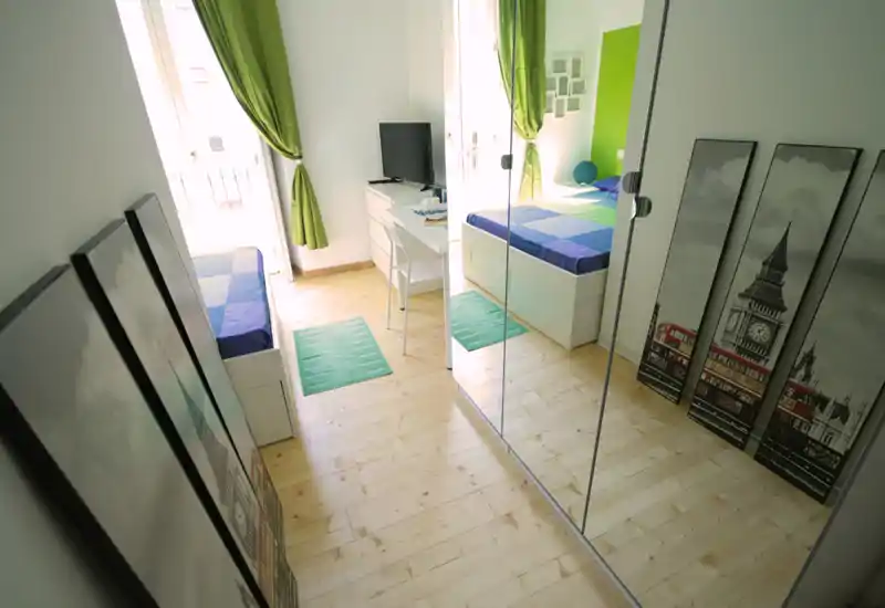 Dormitorio moderno y luminoso con cama individual, escritorio y armario con espejo; acentos verdes y luz natural crean un ambiente acogedor y amplio.