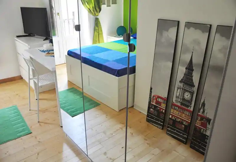 Dormitorio moderno y luminoso con cama colorida, suelo de madera y armario con espejos que aportan profundidad. Luz natural y estilo limpio, perfecto para anuncios de alquiler.