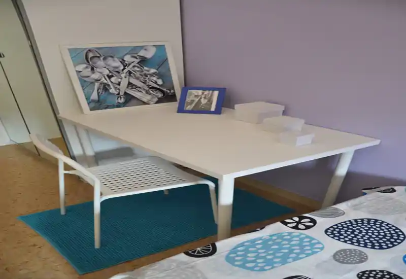 Coin de chambre avec un bureau blanc et une chaise à côté d’un lit à motifs. Décor soigné : cadre photo, petites boîtes de rangement et une affiche appuyée sur le bureau contre un mur violet clair.