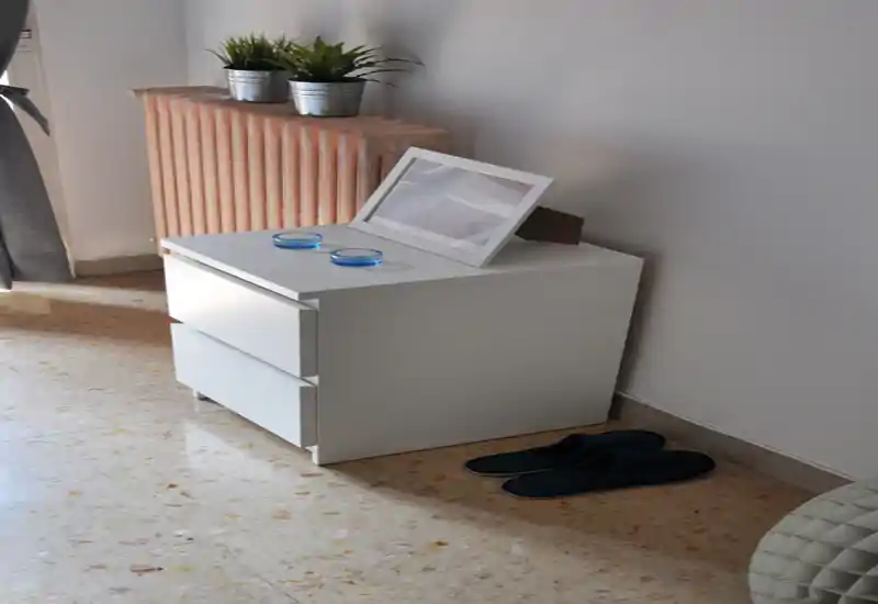 Coin de chambre soigné avec une table de chevet blanche à deux tiroirs, un cadre décoratif, de petites plantes sur le radiateur et des chaussons au sol ; lumineux et minimaliste.