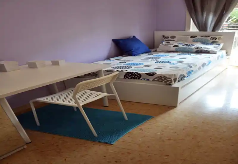 Chambre simple lumineuse avec lit à cadre blanc, couette à motifs et petit bureau à côté. Murs violets clairs, coussin bleu et lumière naturelle venant de la fenêtre.
