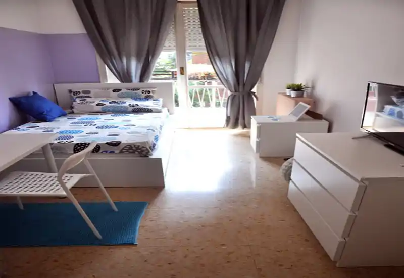 Bright, neatly staged bedroom with a double bed, patterned linens, a small desk and dresser, and direct natural light from a balcony door.