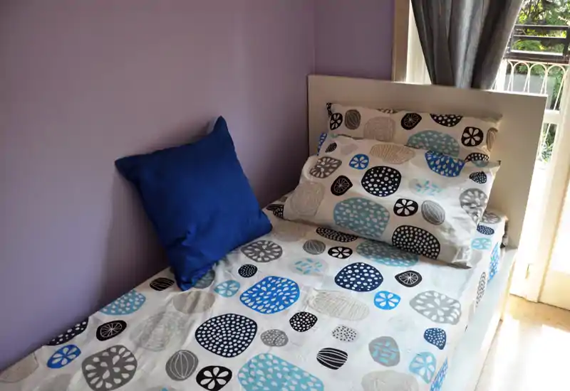 Bright, neatly made single bedroom with patterned bedding and a deep blue accent cushion beside a window letting in natural light.