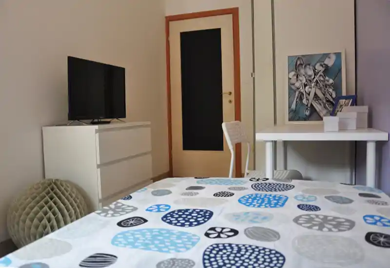 Chambre lumineuse et épurée avec couette à motifs, télévision sur une commode blanche et un petit bureau mural — agencement moderne et fonctionnel, adapté aux locations courtes.