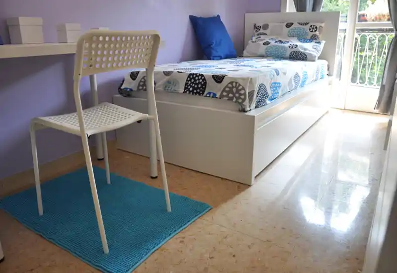 Bright single bedroom with a white bed frame, patterned bedding and a small workspace. Natural light enters from a balcony door, creating a clean, welcoming atmosphere ideal for listings.