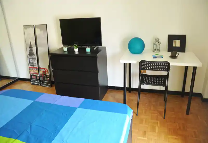 Bright, simple bedroom with a neatly made bed in blue tones, a dresser with TV and a small desk ideal for work or study.