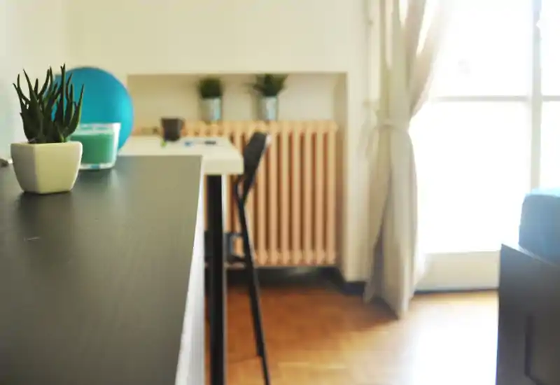 Bright, casual living area corner with a small table, stool and decorative plants; warm wooden floor and natural light from a large window create a relaxed atmosphere.