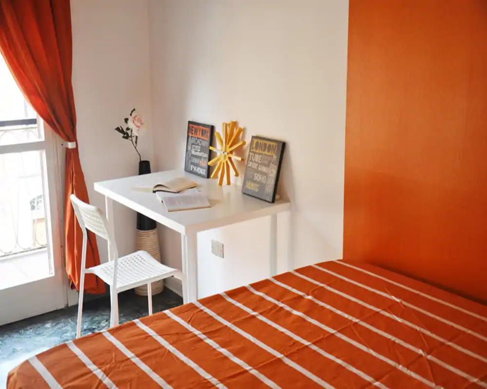 Bright, warmly decorated bedroom with an orange-striped bedspread, a small white desk by the window and decorative accents that create a cozy workspace.