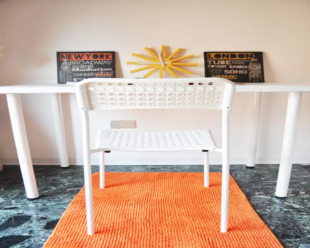Bright, minimalist workspace with a white perforated chair, slim desk, orange rug and decorative wall art — a compact, cheerful study corner.