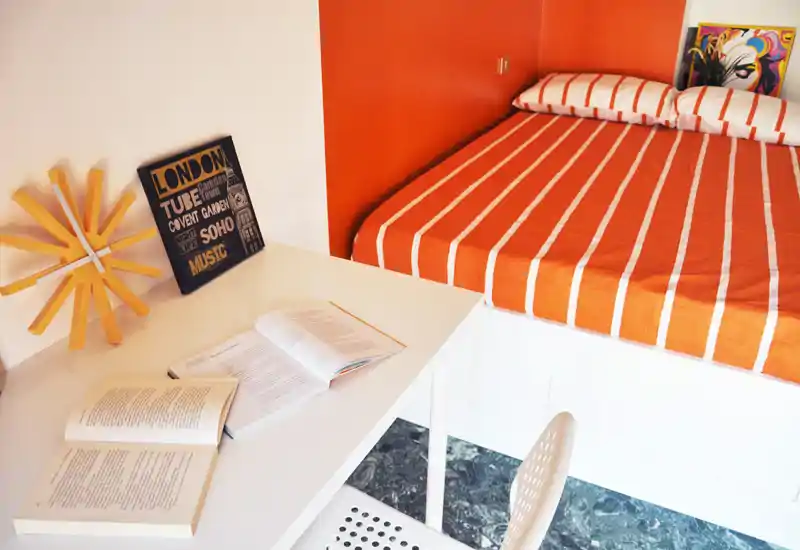 Bright, modern bedroom corner with an orange-striped bed and a tidy white desk with open books — ideal for a compact, styled studio.