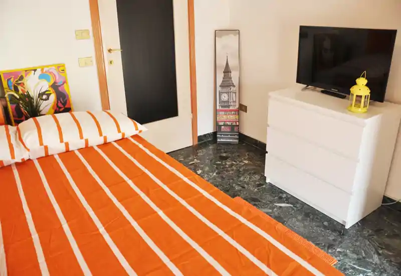 Bright, neatly styled bedroom with an orange-striped bedspread, decorative artwork and a white chest of drawers with TV—pleasant and welcoming sleeping space.