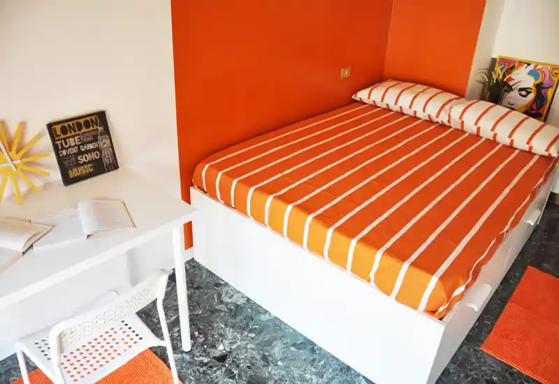 Compact, well-styled bedroom with an orange-striped double bed, matching textiles and a small desk — bright and inviting for a solo or couple stay.