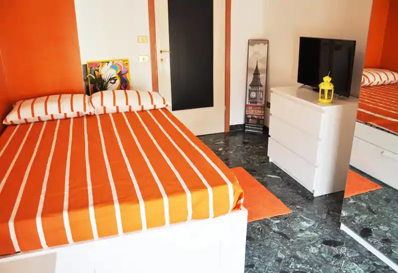 Bright, modern bedroom with an orange striped double bed, matching textiles and a white dresser with TV. The room features warm accents and a large mirror that enhances perceived space.