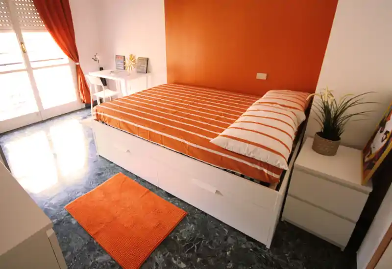 Bright, modern bedroom with an orange-accented double bed, coordinated linens and a small desk by the window. Large window/door provides abundant natural light and a cozy rug adds warmth.