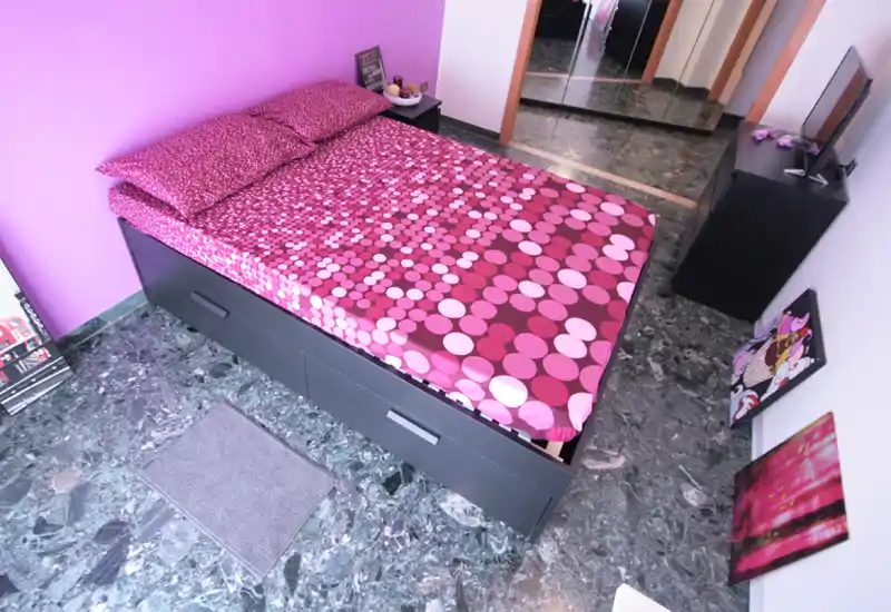 Bright, well-styled bedroom with a patterned pink double bed, bedside table and mirrored wardrobe; bold purple accent wall and marble floor create a modern, vibrant look.