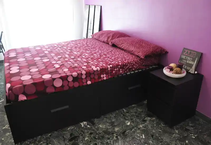 Bright, stylish bedroom with a patterned double bed against a purple accent wall and a black bedside table. Natural light filters from the window, and coordinated linens give the room a modern, cozy feel ideal for rental photography.