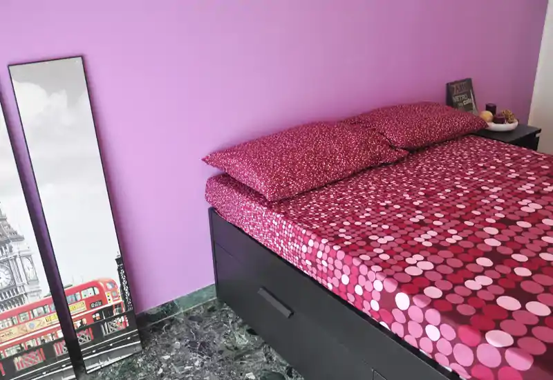 Bright, styled bedroom with a patterned double bed against a pastel pink wall, bedside table and decorative mirror — a cozy, well-lit sleeping space.
