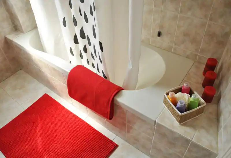 Bright, clean bathroom with a curved bathtub, white shower curtain with black teardrop pattern, red bath mat and towel, and toiletries in a small basket.