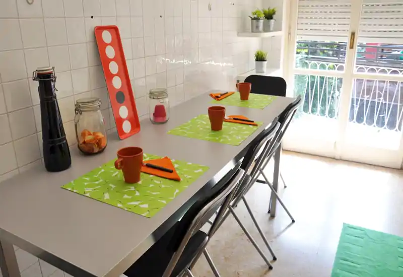 Bright, modern dining area with a long table set for three, colorful placemats and cups, and natural light from a balcony door — ideal for casual meals.