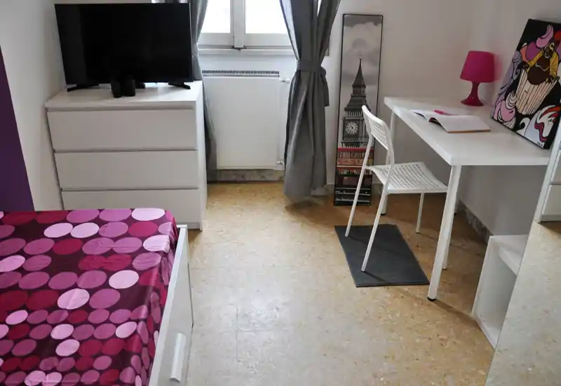 Chambre compacte et lumineuse avec lit à motifs, commode blanche avec télévision et petit bureau près de la fenêtre — sobre, moderne et fonctionnelle.