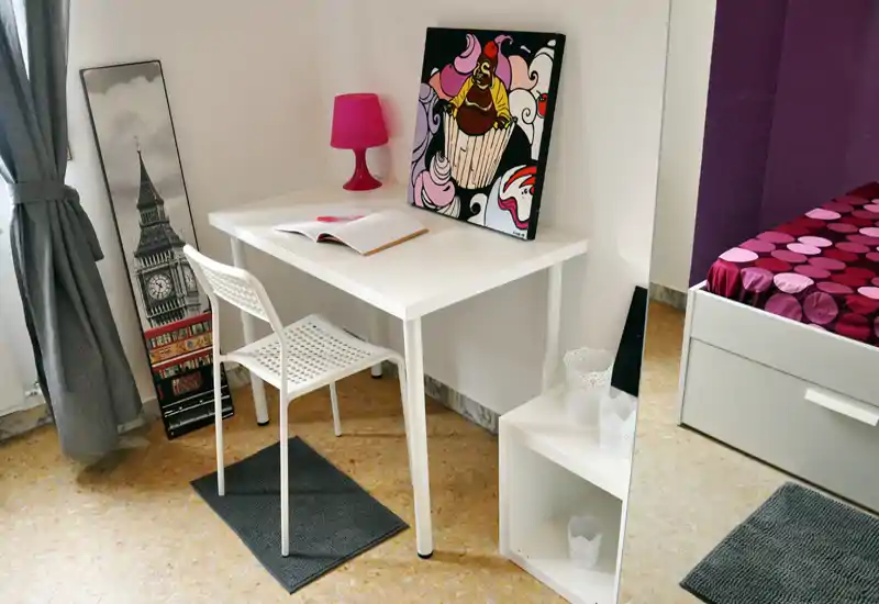 Coin de chambre lumineux et soigné avec un bureau blanc, une chaise et un lit partiellement visible avec une literie colorée ; parfait pour étudier et se détendre.