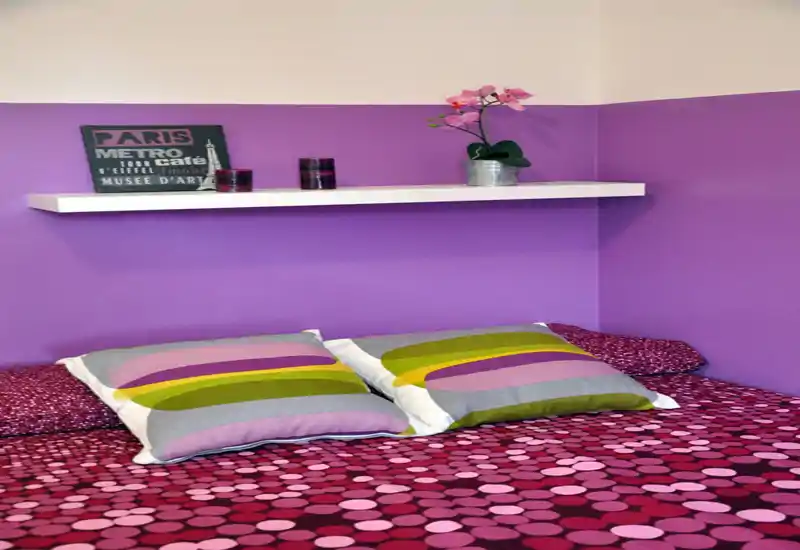 Coin chambre lumineux et coloré avec une couette à motifs, deux coussins décoratifs et une étagère flottante avec petits objets—chaleureux et bien mis en scène.