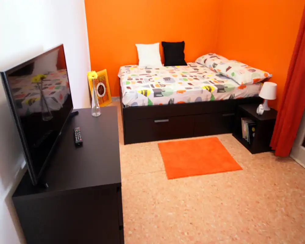 Compact bedroom with a single bed set against a bright orange accent wall, bedside table with lamp, a small TV unit and a matching orange rug — a cheerful, well-lit space ideal for one person.