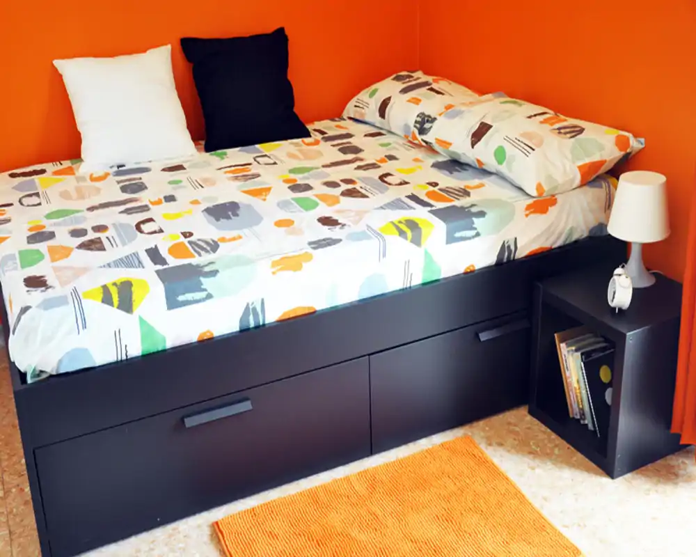 Sunny, compact bedroom with a full view of the bed, colorful bedding and warm orange walls — cozy and modern micro-bedroom ideal for a single occupant.