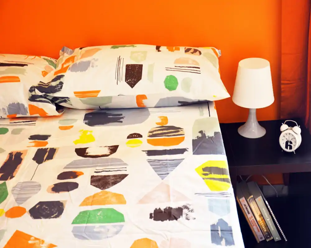 Bright, colorful bedroom corner with patterned bedding, pillows and a bedside table with lamp and alarm clock—warm orange wall adds a cozy atmosphere.