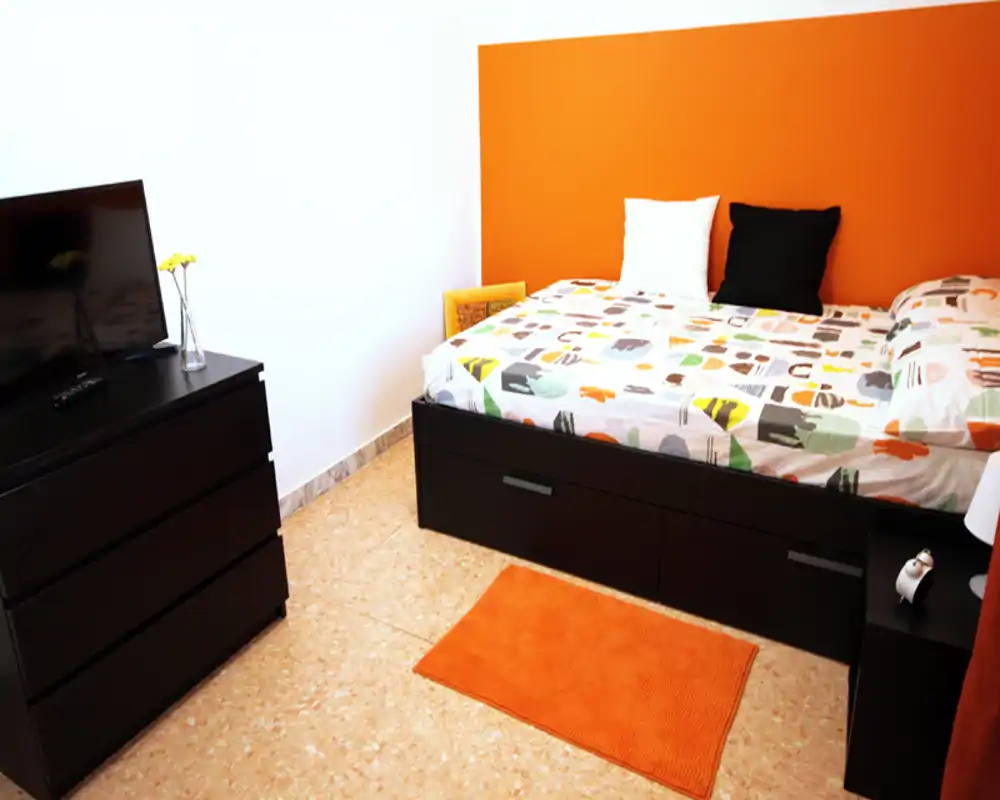 Sunny, compact bedroom with an orange accent wall, patterned bedding and a matching rug. Dark wood storage under the bed and a dresser provide practical space for stays.