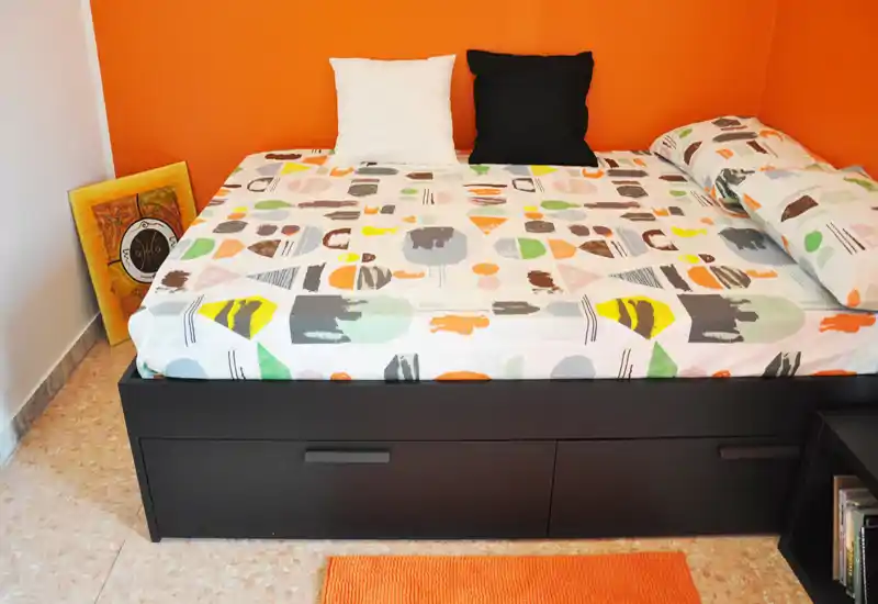 Chambre lumineuse et moderne avec un grand lit plateforme devant un mur orange vif, linge de lit à motifs et deux coussins; sol carrelé et petit tapis orange complètent la pièce.