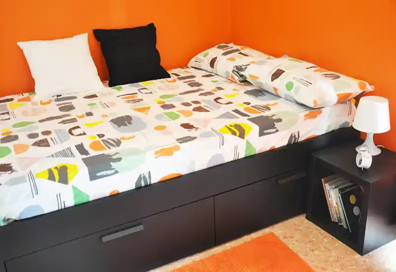 Chambre simple moderne et lumineuse avec couette à motifs, lit coffre et table de chevet avec lampe — le mur orange apporte une ambiance chaleureuse.