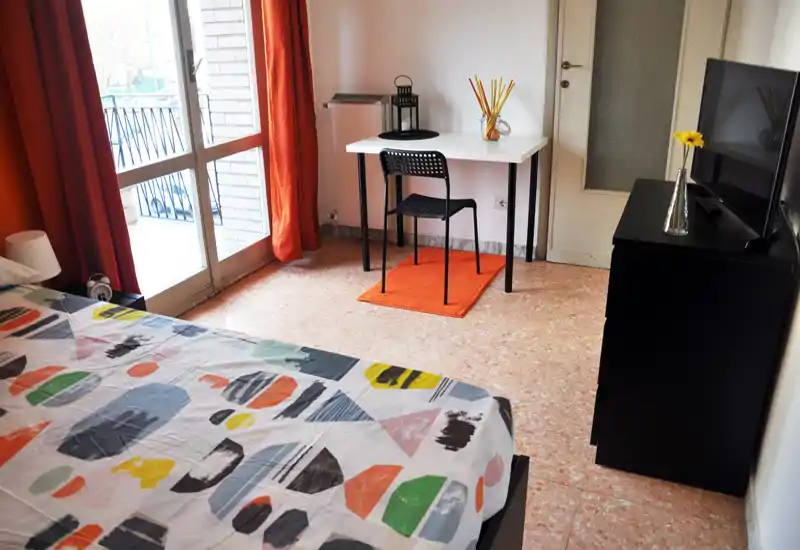 Chambre lumineuse et colorée avec couette à motifs, porte-fenêtre donnant sur balcon et un petit bureau — aérée et bien éclairée, idéale pour un séjour confortable.