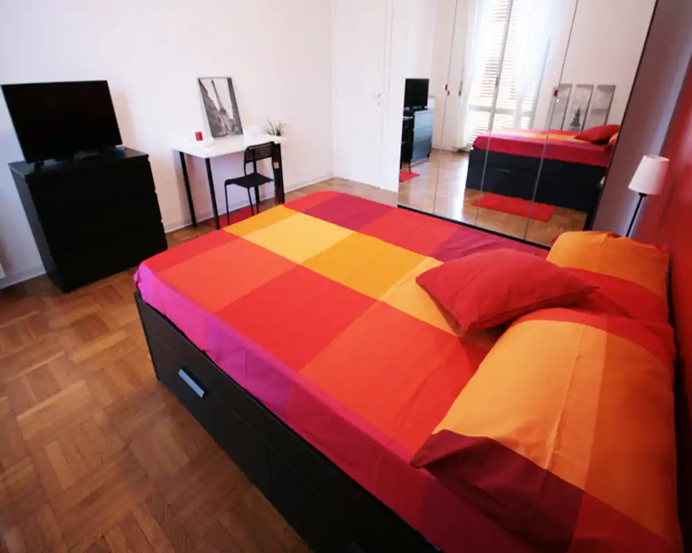 Camera da letto luminosa e ben composta con un ampio letto matrimoniale colorato, armadio con specchi, TV e una piccola scrivania — ideale per un soggiorno vivace in città.