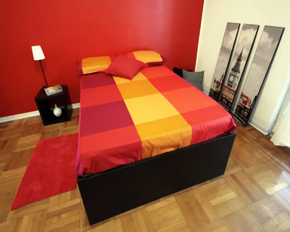 Camera da letto luminosa e vivace con letto matrimoniale rifinito con lenzuola rosso-arancio, lampada da comodino, pavimento in parquet e quadri decorativi a parete.
