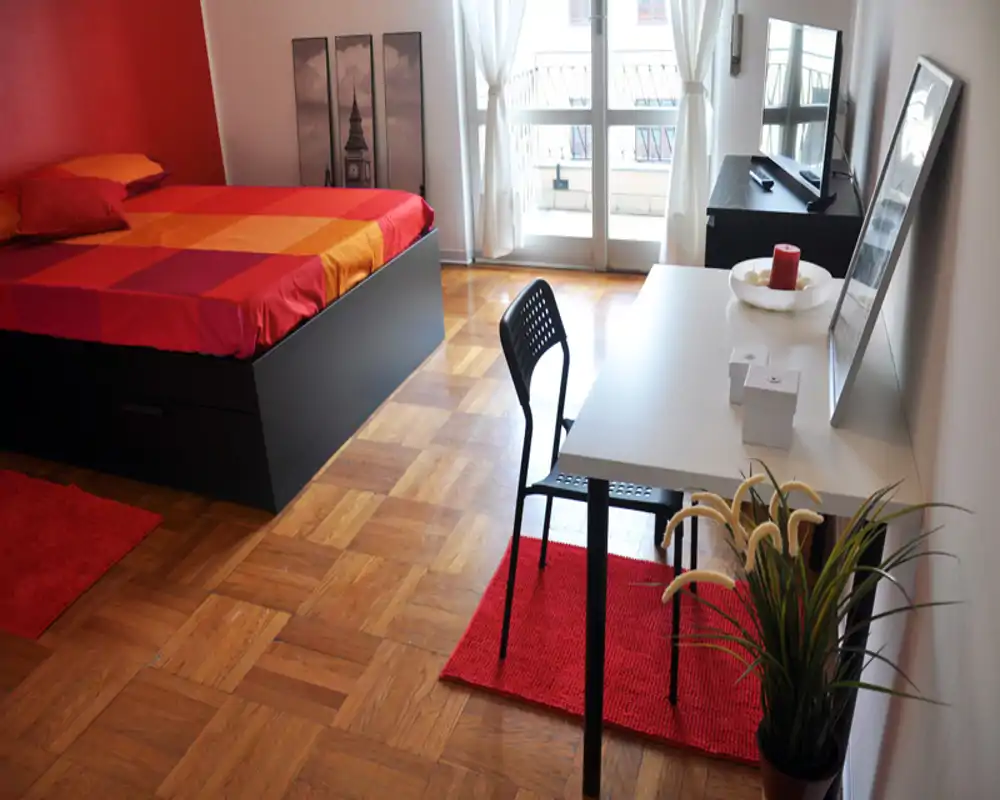 Camera da letto luminosa e ben arredata con grande letto contenitore, tessuti rossi coordinati e una scrivania vicino alla finestra con accesso al balcone.