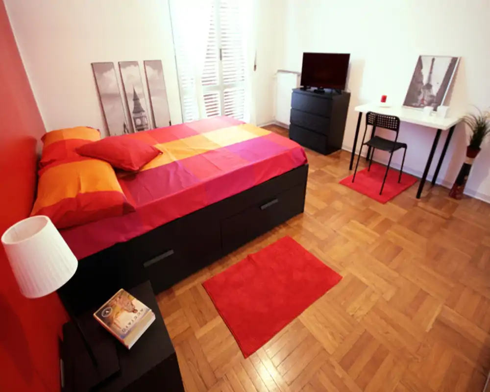 Camera da letto moderna e luminosa con un grande letto, biancheria rossa e arancione, pavimento in parquet e una piccola scrivania. La stanza è ben illuminata dalla finestra con persiane e arredata in stile contemporaneo, ideale per una persona o una coppia.