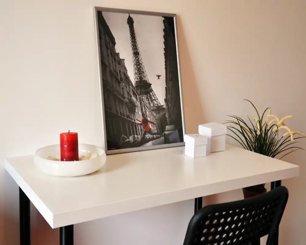 Piccolo angolo con scrivania dallo stile moderno: stampa incorniciata di Parigi, candela rossa e pianta in vaso; ideale come postazione compatta o elemento decorativo.