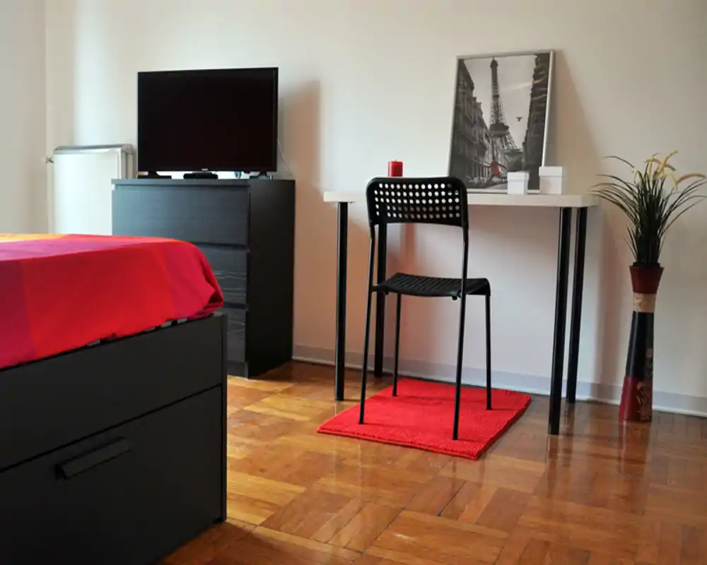 Camera da letto luminosa e moderna con letto nero e biancheria rossa, una scrivania con sedia e una TV su comò; pavimento in parquet e arredo minimal.